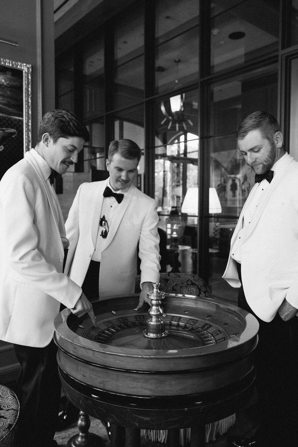 Groomsmen in white dinner jackets gathered around a bar in black and white