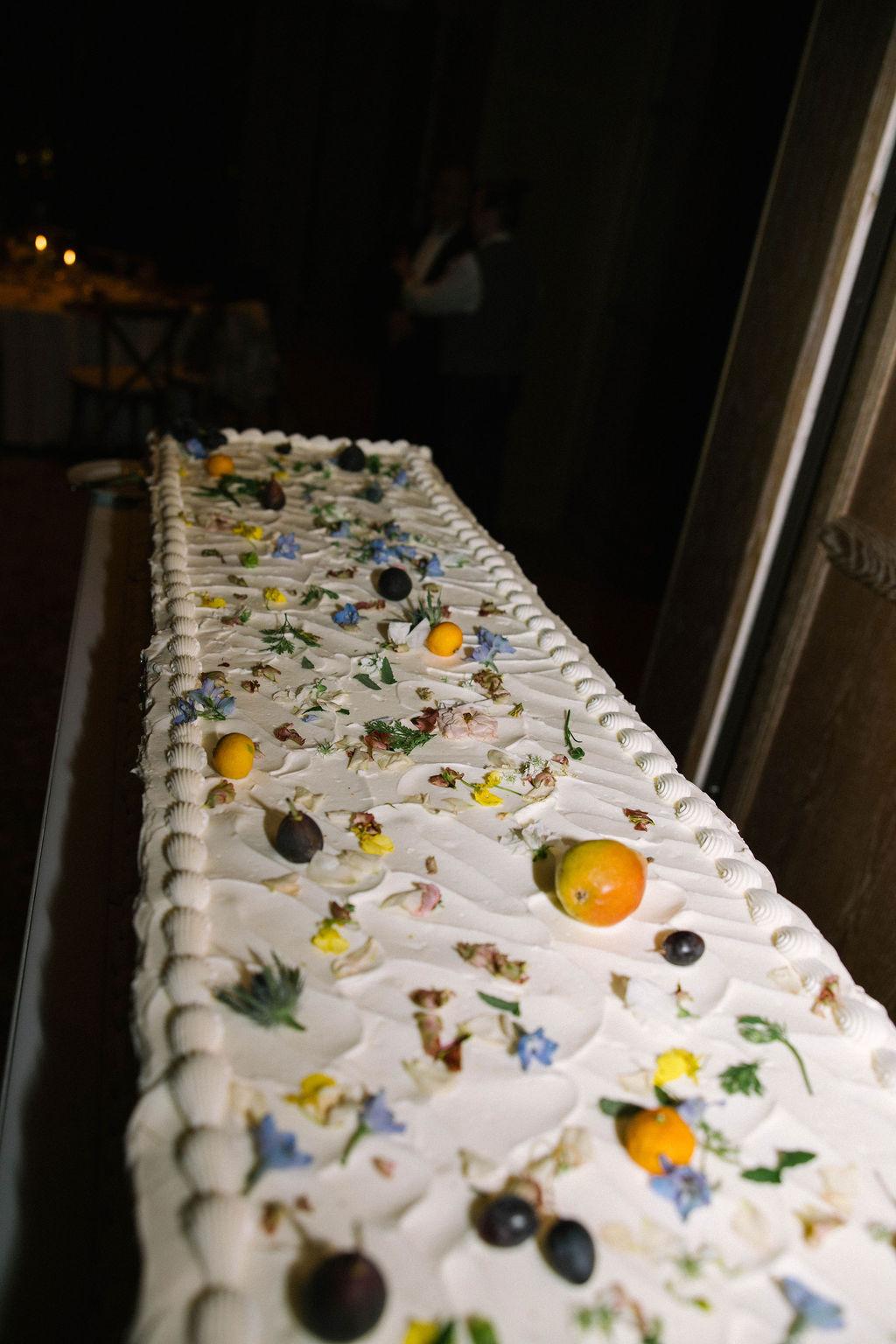Long banquet table dressed with a runner strewn with fruit and blossoms at night
