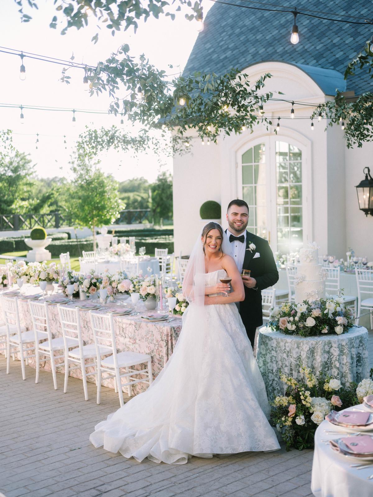 A couple stand with drinks in front of a long reception table dressed in floral linens