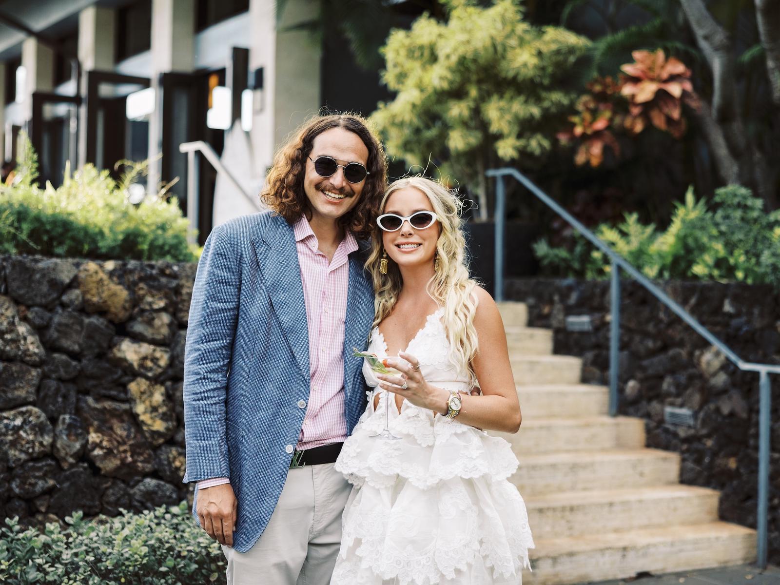 A couple in sunglasses stand together on resort steps surrounded by tropical greenery