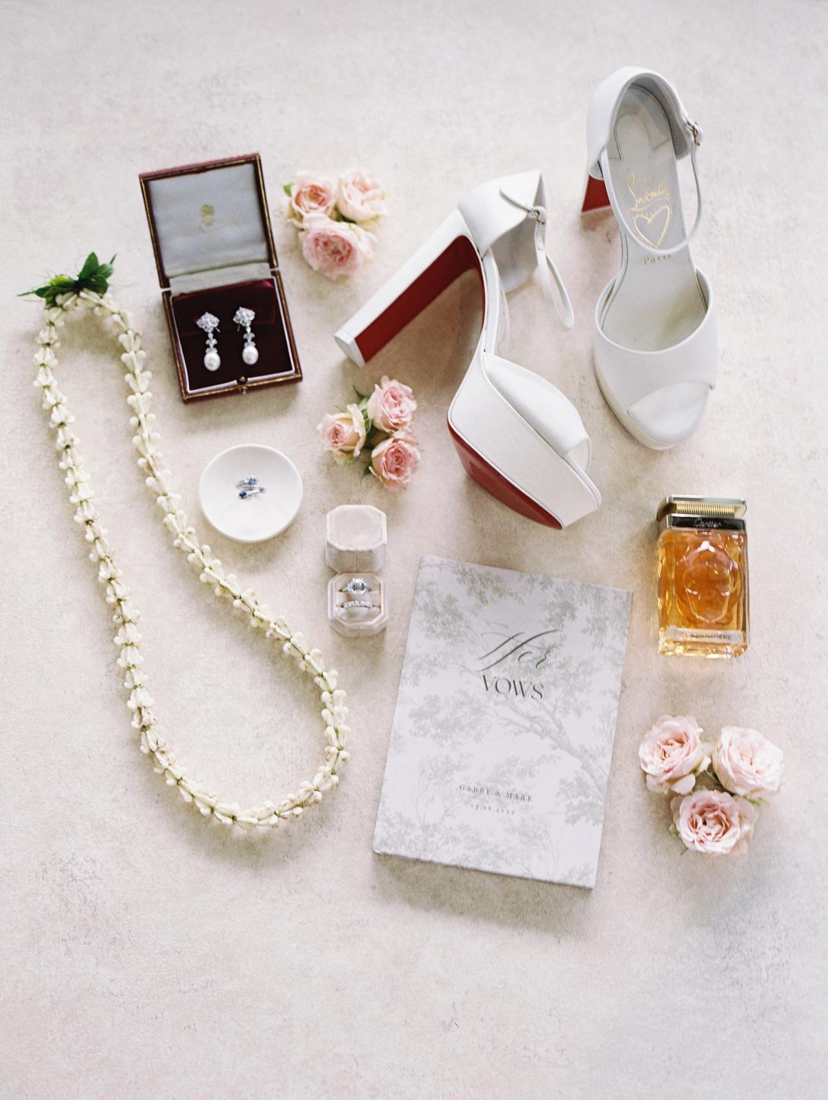A flat lay of bridal shoes, a pearl necklace, perfume, earrings and the invitation suite