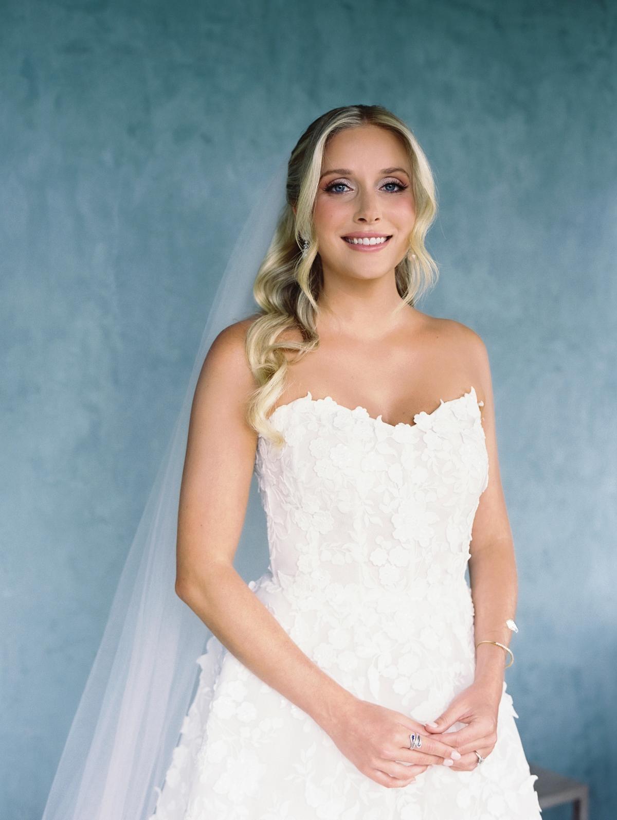 A portrait of the bride in a strapless lace gown and veil against a blue-green backdrop