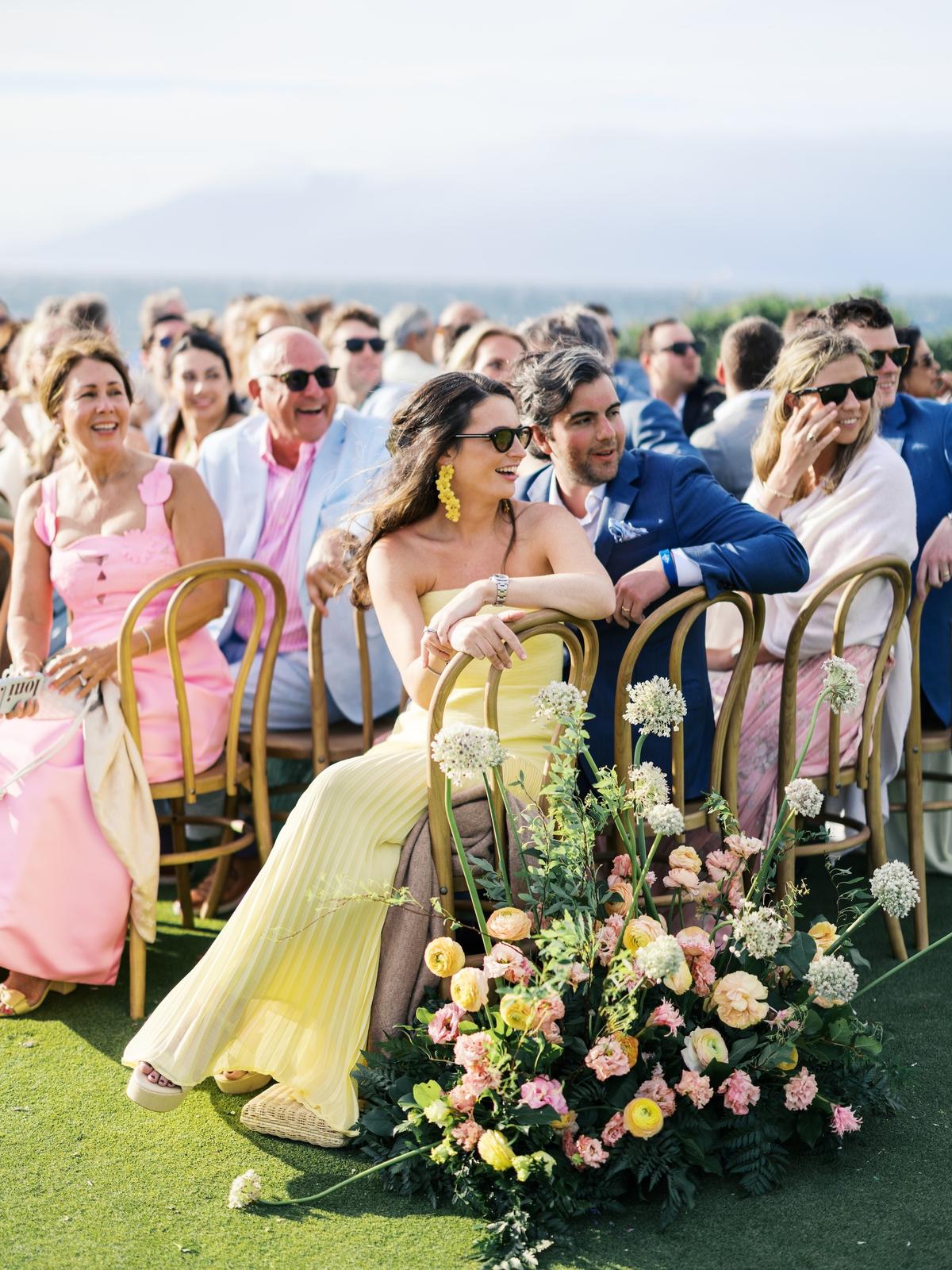 Wedding guests in pastel dresses seated on cane chairs at an outdoor ceremony