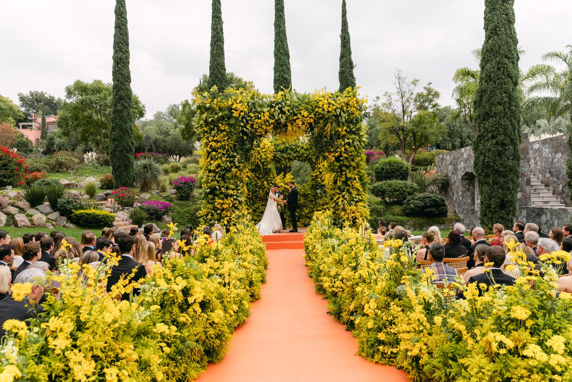 A garden ceremony aisle lined with yellow blooms and cypress trees, with an orange runner leading to a floral arch