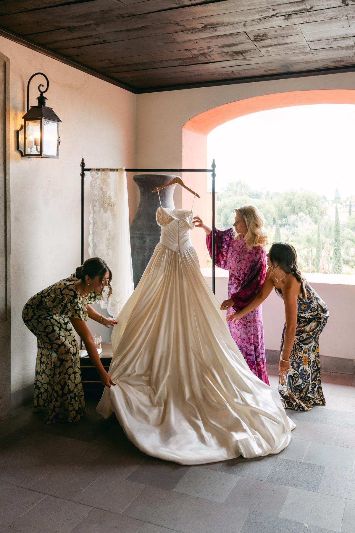 Three attendants in printed dresses steam and arrange a full ivory wedding gown on a hanger