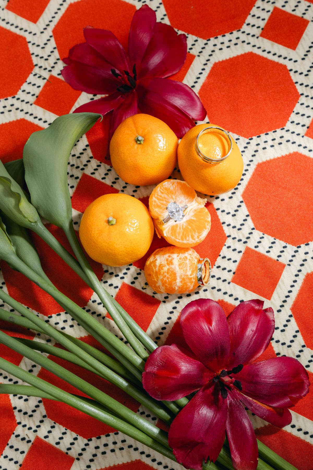 Overhead flat lay of citrus fruit, burgundy tulips and calla lily stems on a patterned orange textile