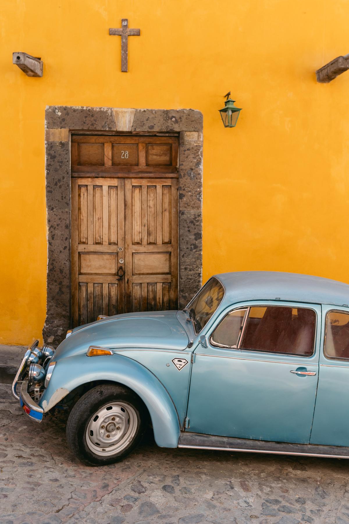 A vintage pale blue car parked against a saturated yellow wall with a carved wooden door