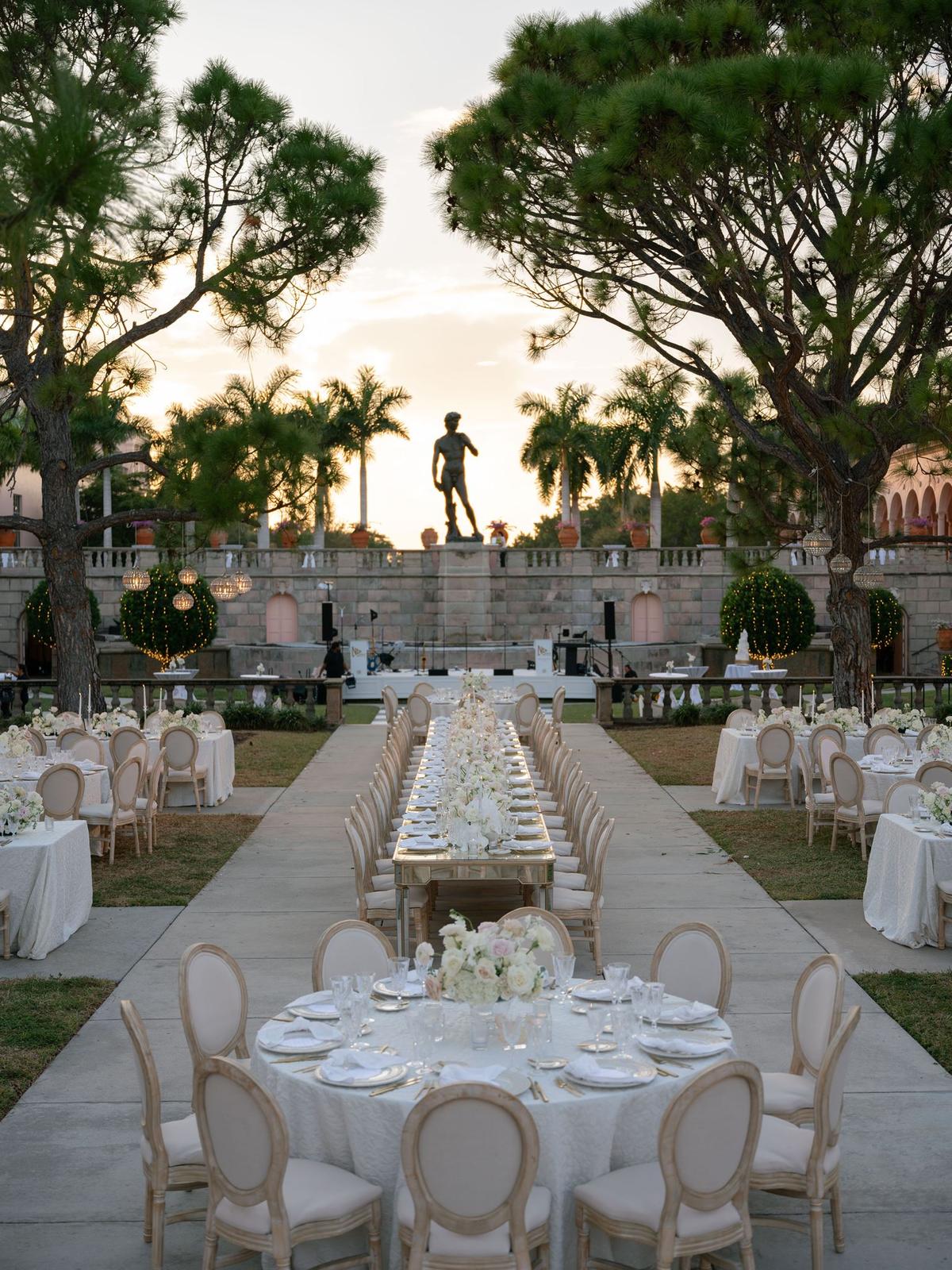 Long banquet tables set with white linens and low florals in a courtyard at dusk