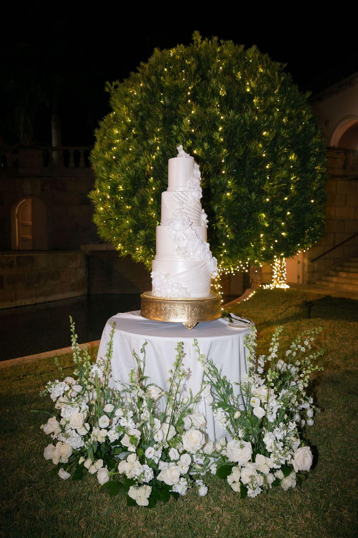 Tiered white wedding cake on a gold stand in front of a lit topiary at night