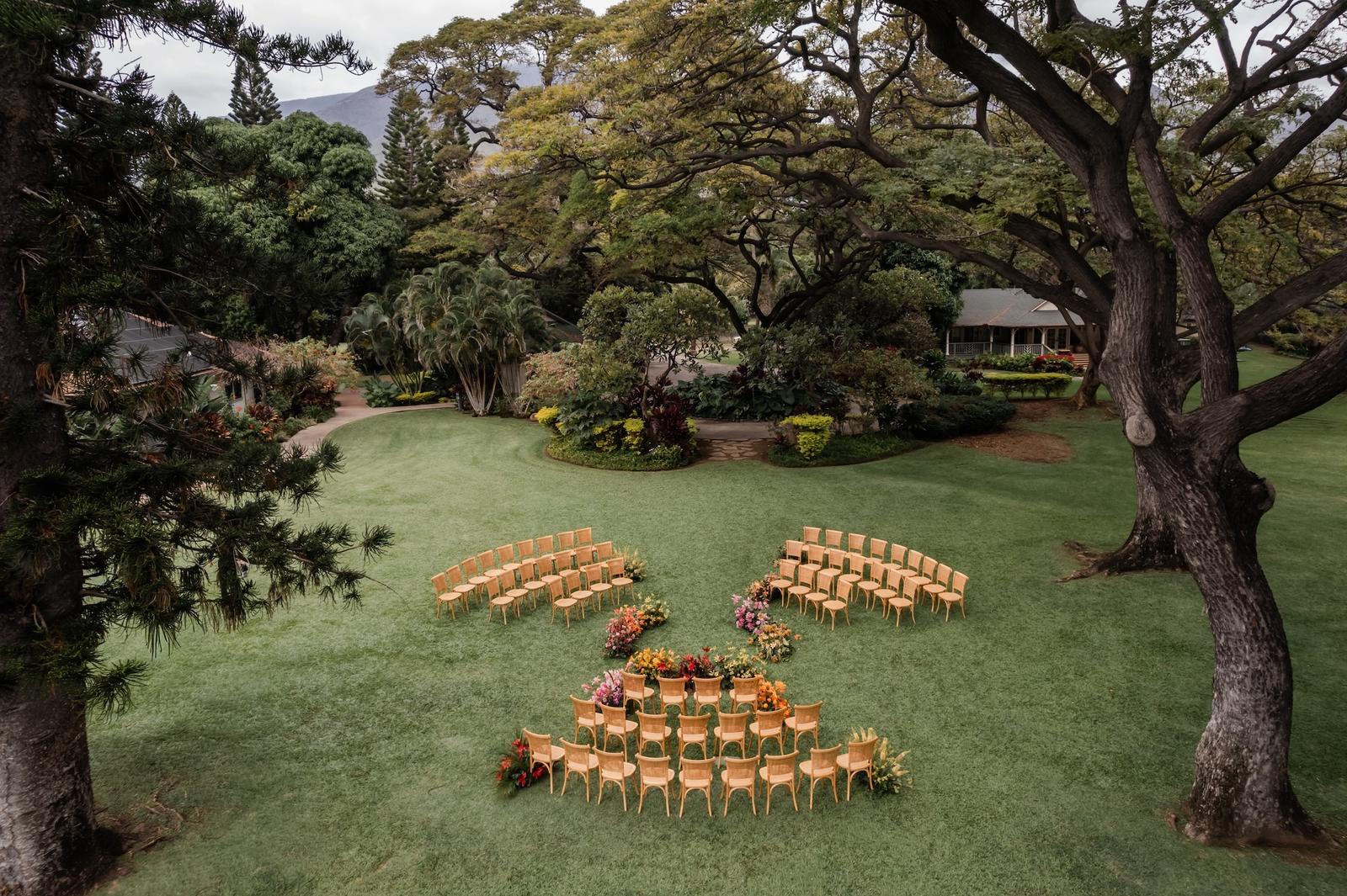 Ceremony chairs arranged under large trees on a lawn in Maui
