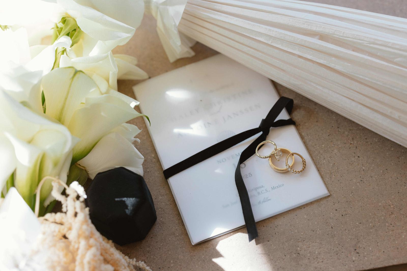 Wedding rings resting on an invitation tied with black ribbon, beside white calla lilies