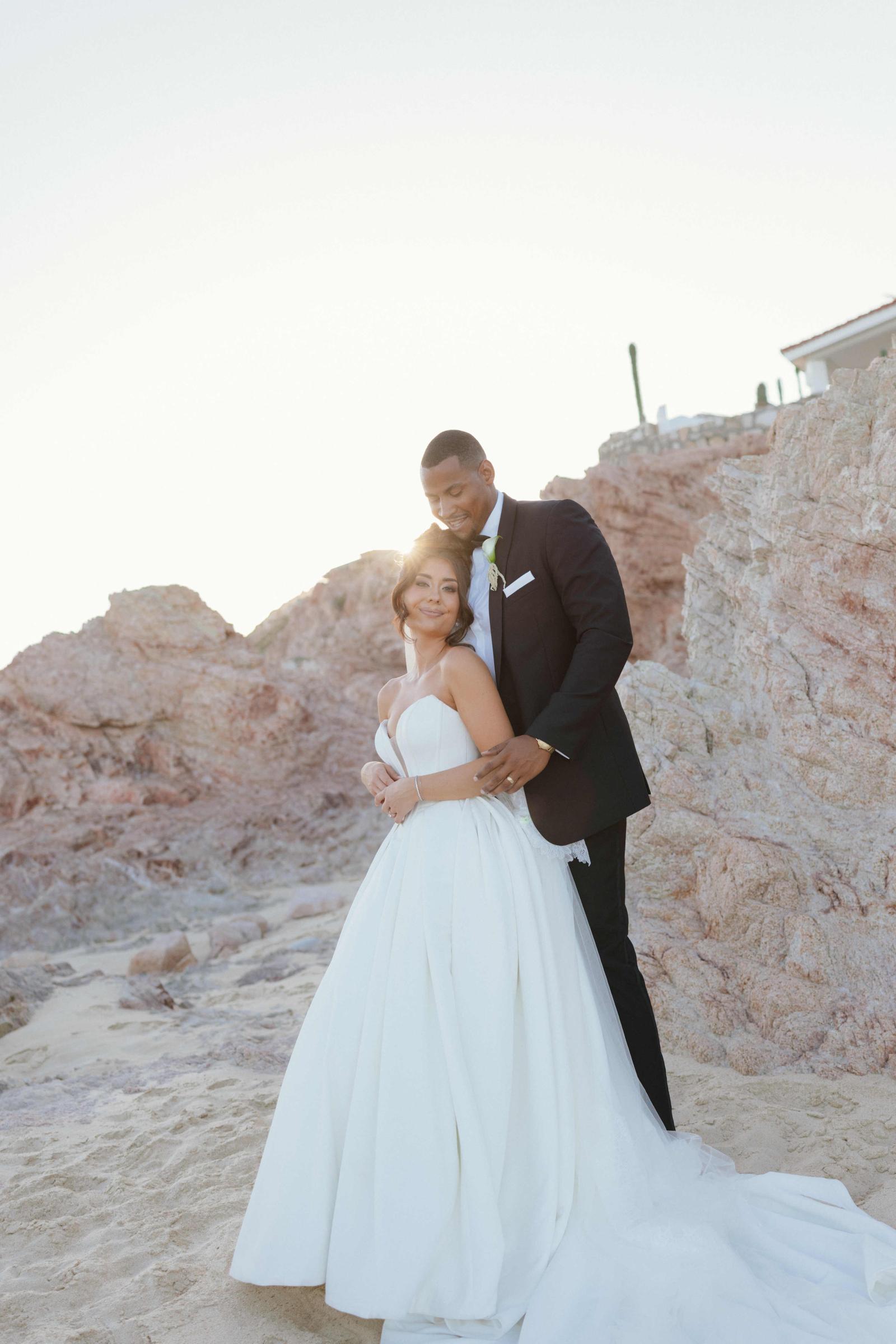 The couple embrace on the sand in front of pink rock formations