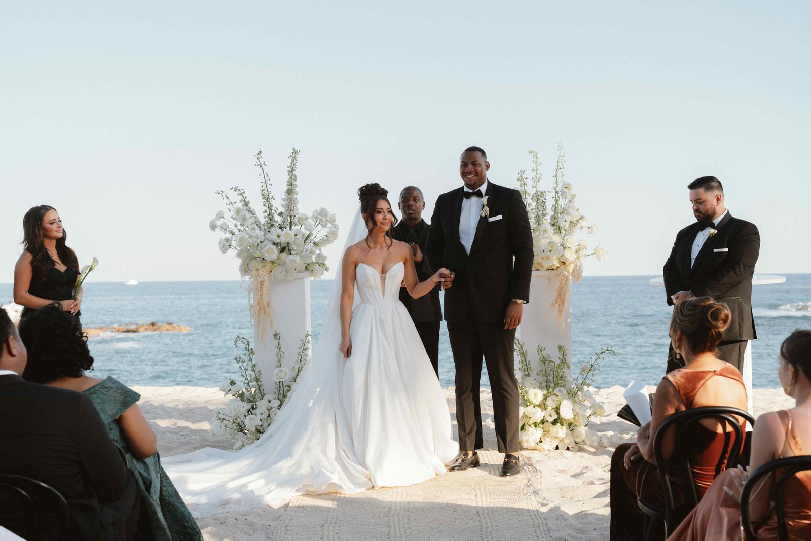 A couple stand at their beachfront ceremony in Los Cabos, framed by white flowers with the ocean behind them