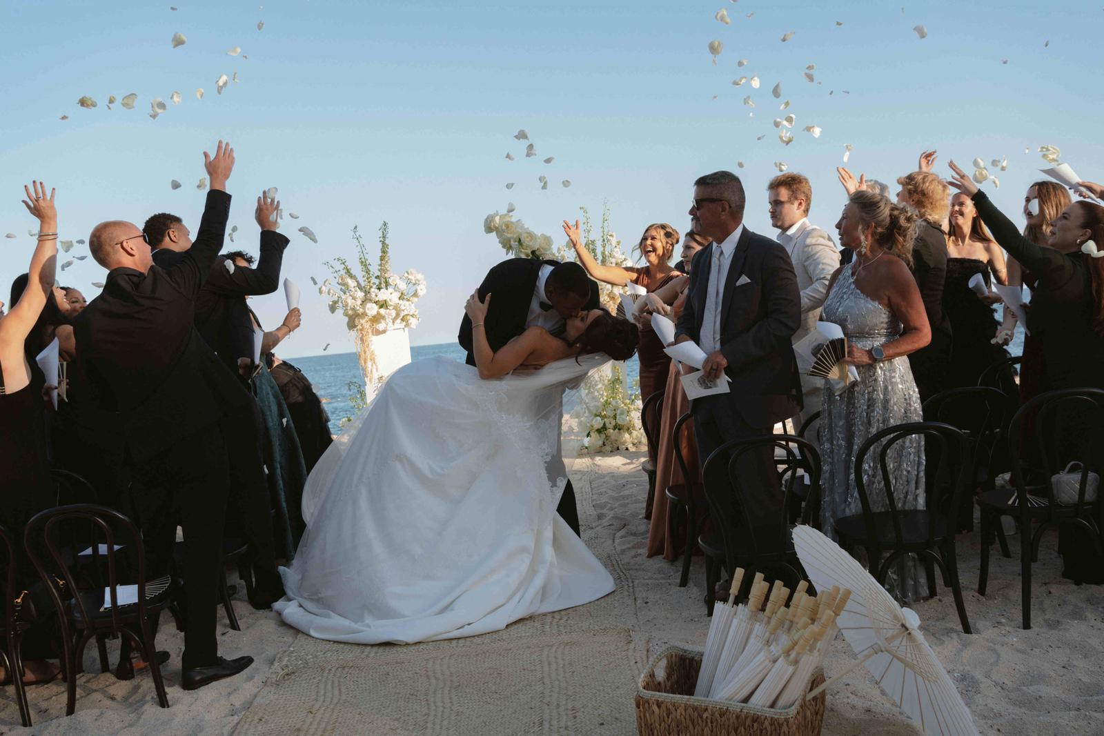 Guests throw petals as the couple kiss at the end of their beach ceremony