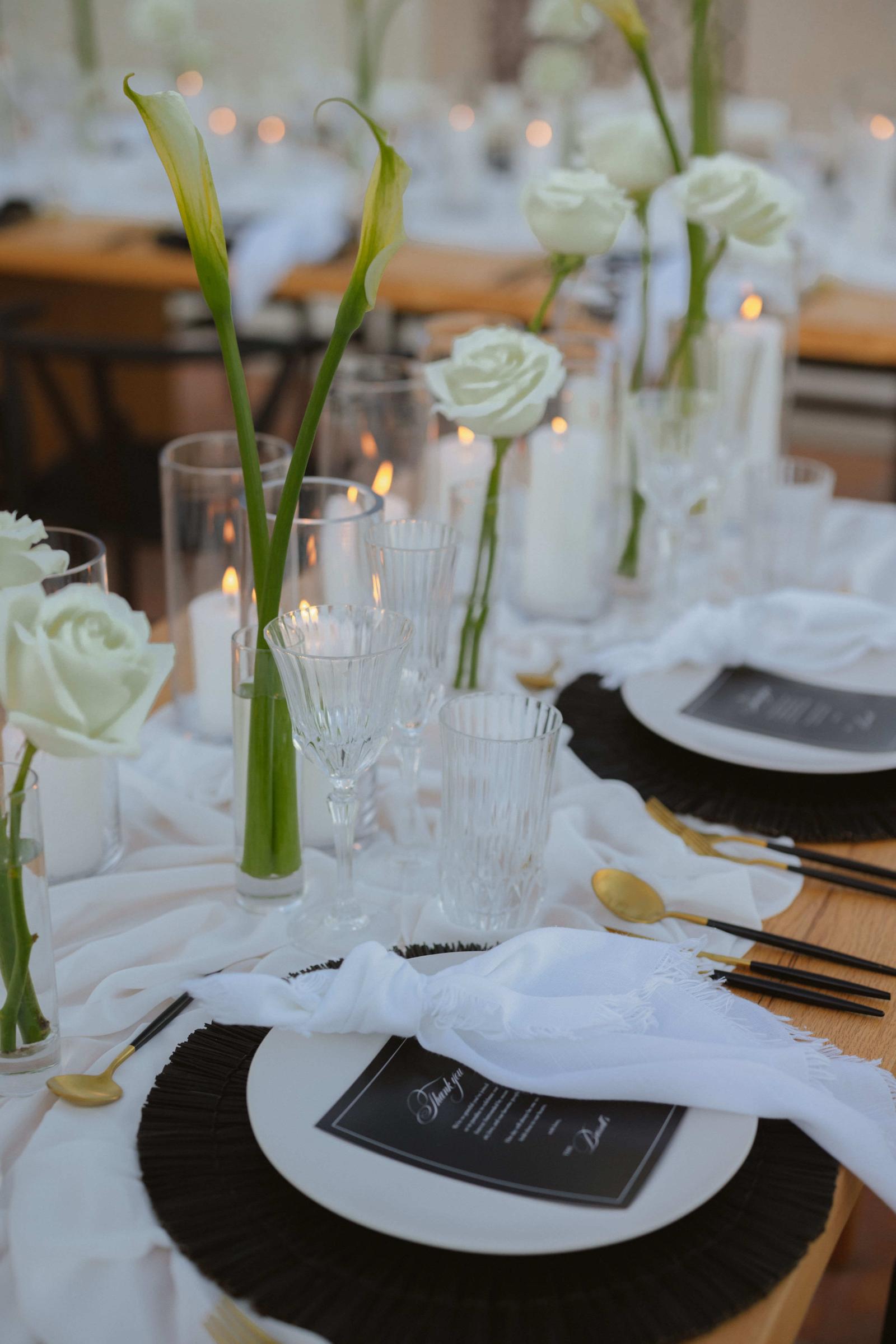 A place setting with a black charger, a white napkin and calla lilies in bud vases