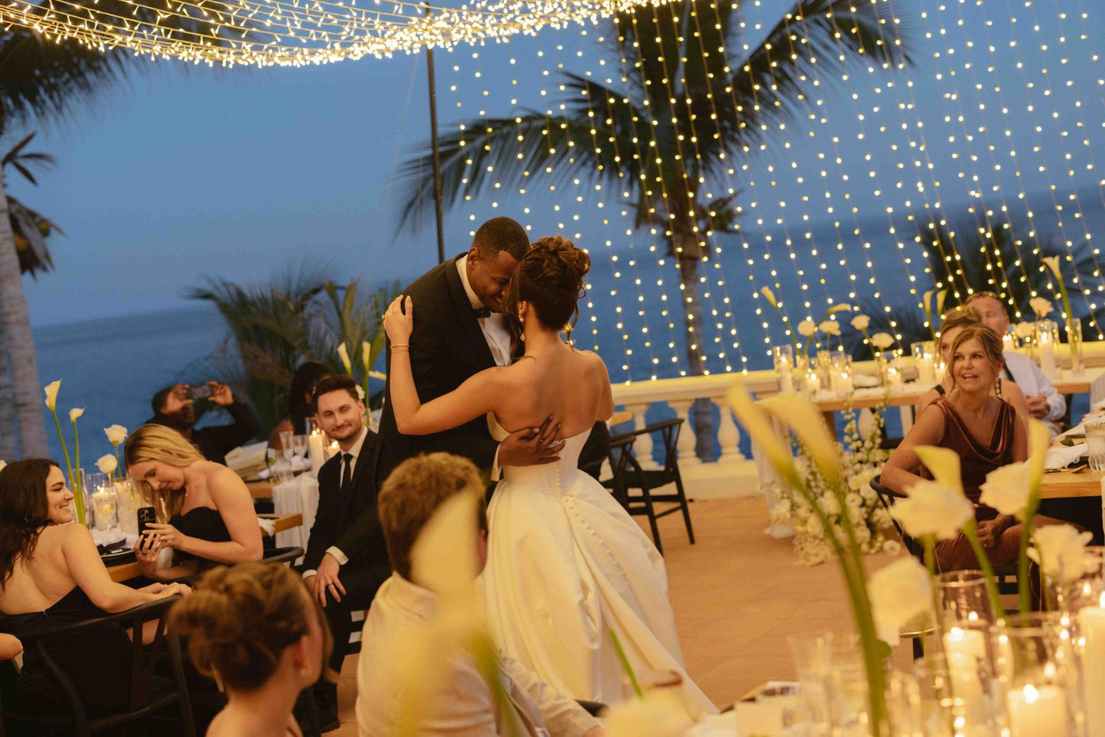 The couple dance at dusk under string lights while seated guests look on