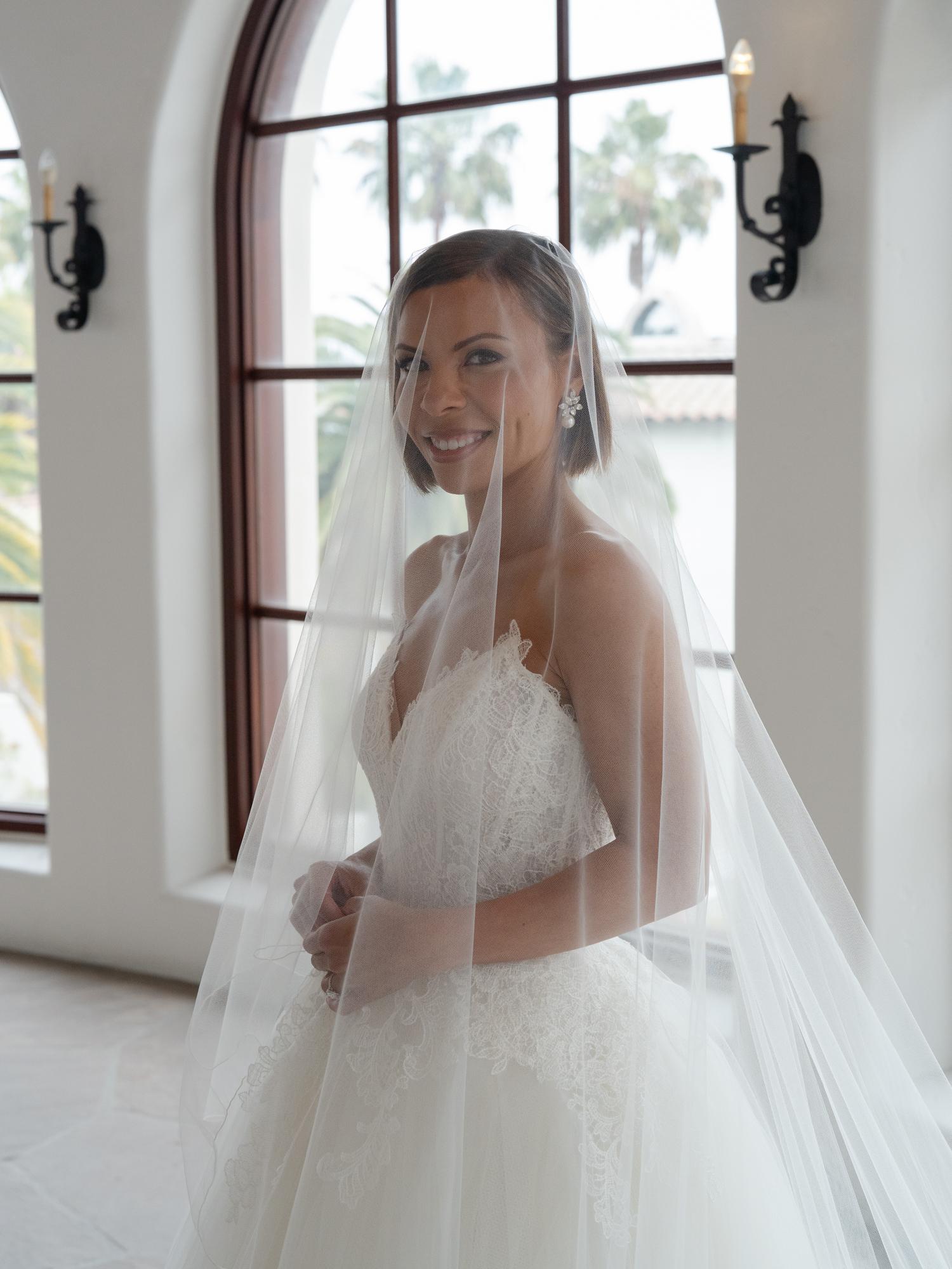 A bride in a lace gown and long veil smiles beside an arched window