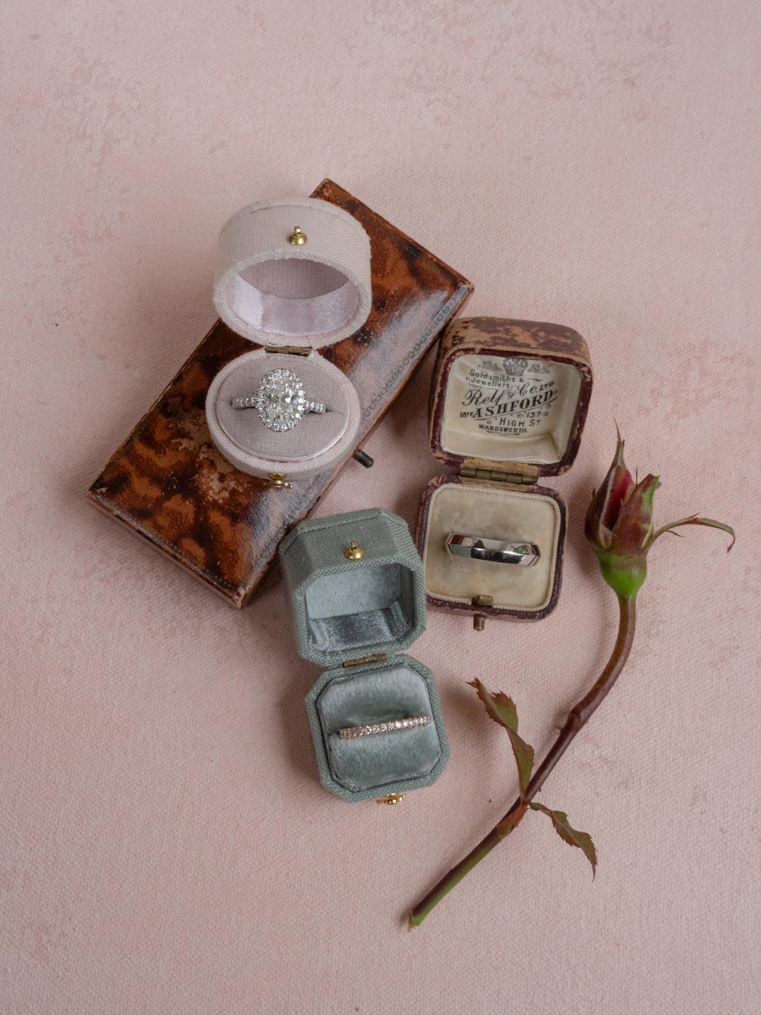 Three vintage ring boxes holding an engagement ring and wedding bands on a pink surface