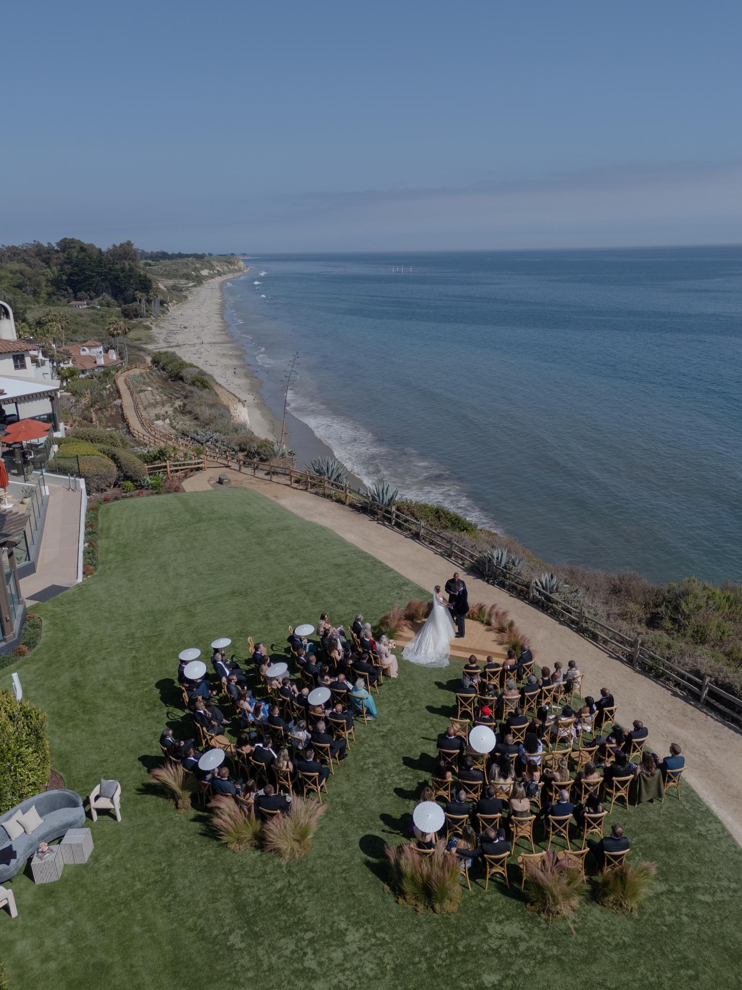 Aerial view of a wedding ceremony on a lawn at the edge of a bluff above the ocean