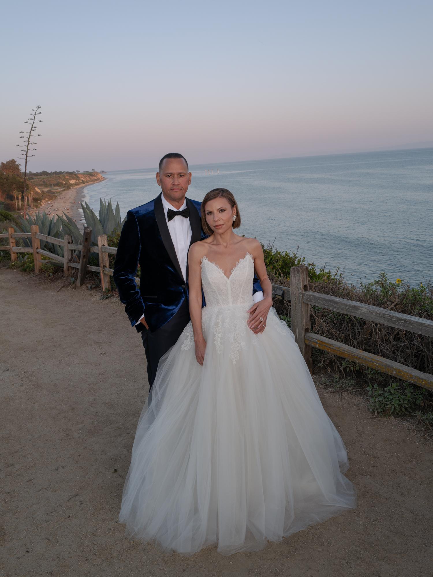 The couple stand together on a bluff path at dusk with the ocean behind them